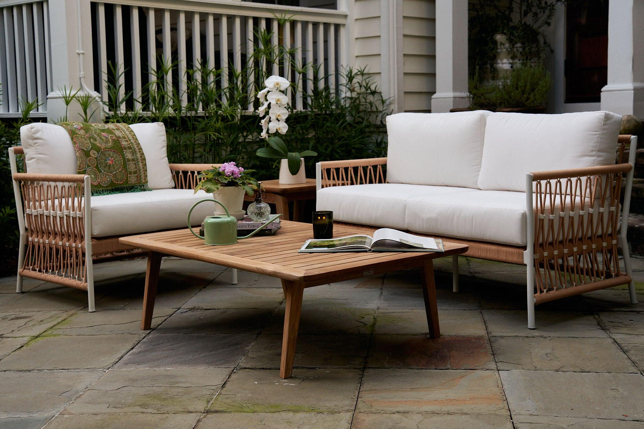 Modern outdoor patio seating area featuring white cushioned sofas with woven wooden frames, a matching wooden coffee table, decorative plants, and cozy contemporary garden décor on a stone terrace.