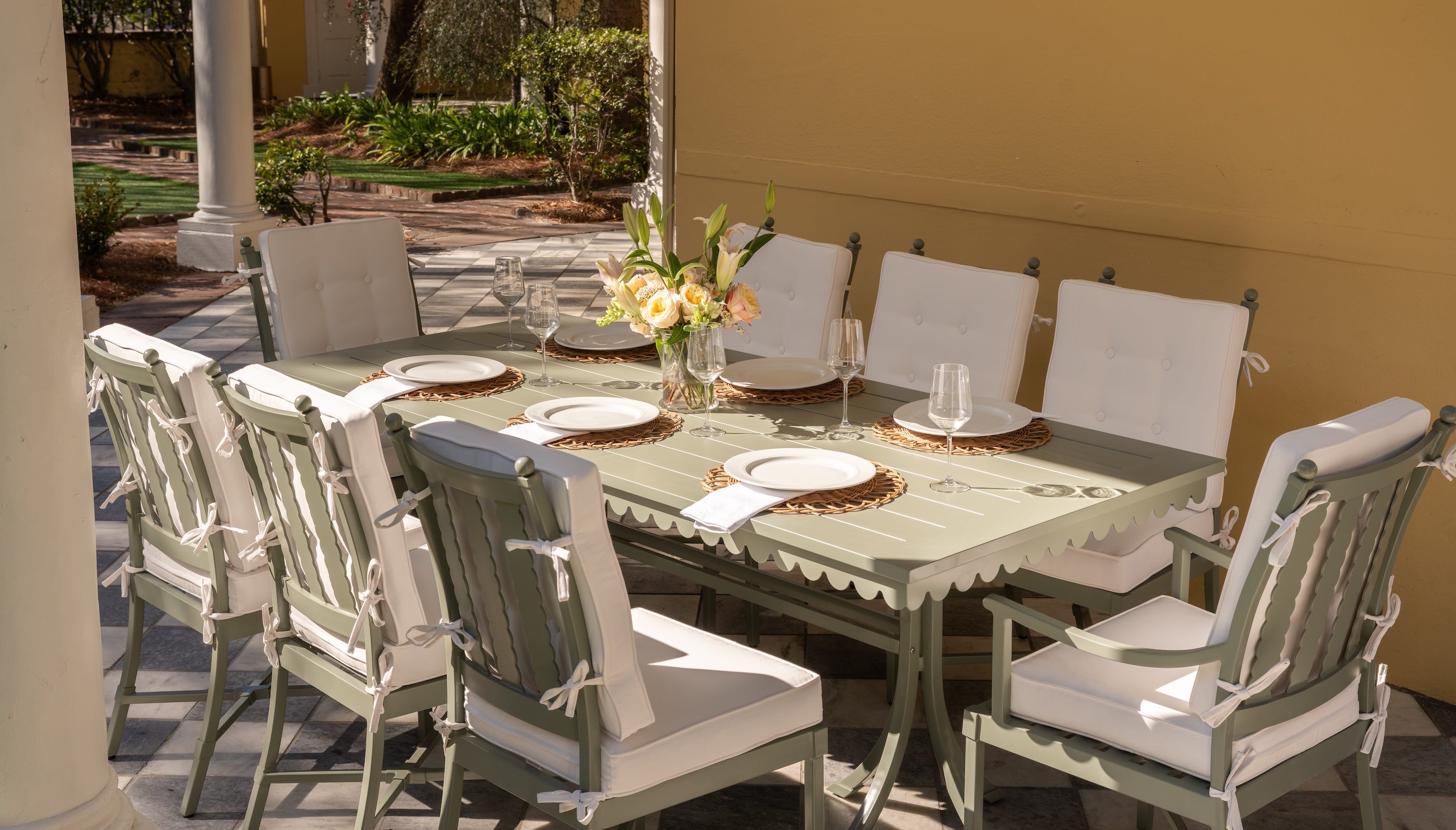 Outdoor dining area featuring a rectangular metal table with a decorative table runner, white cushioned chairs, elegant place settings, and a vase of fresh flowers, set on a patio with a warm yellow wall and lush greenery in the background.
